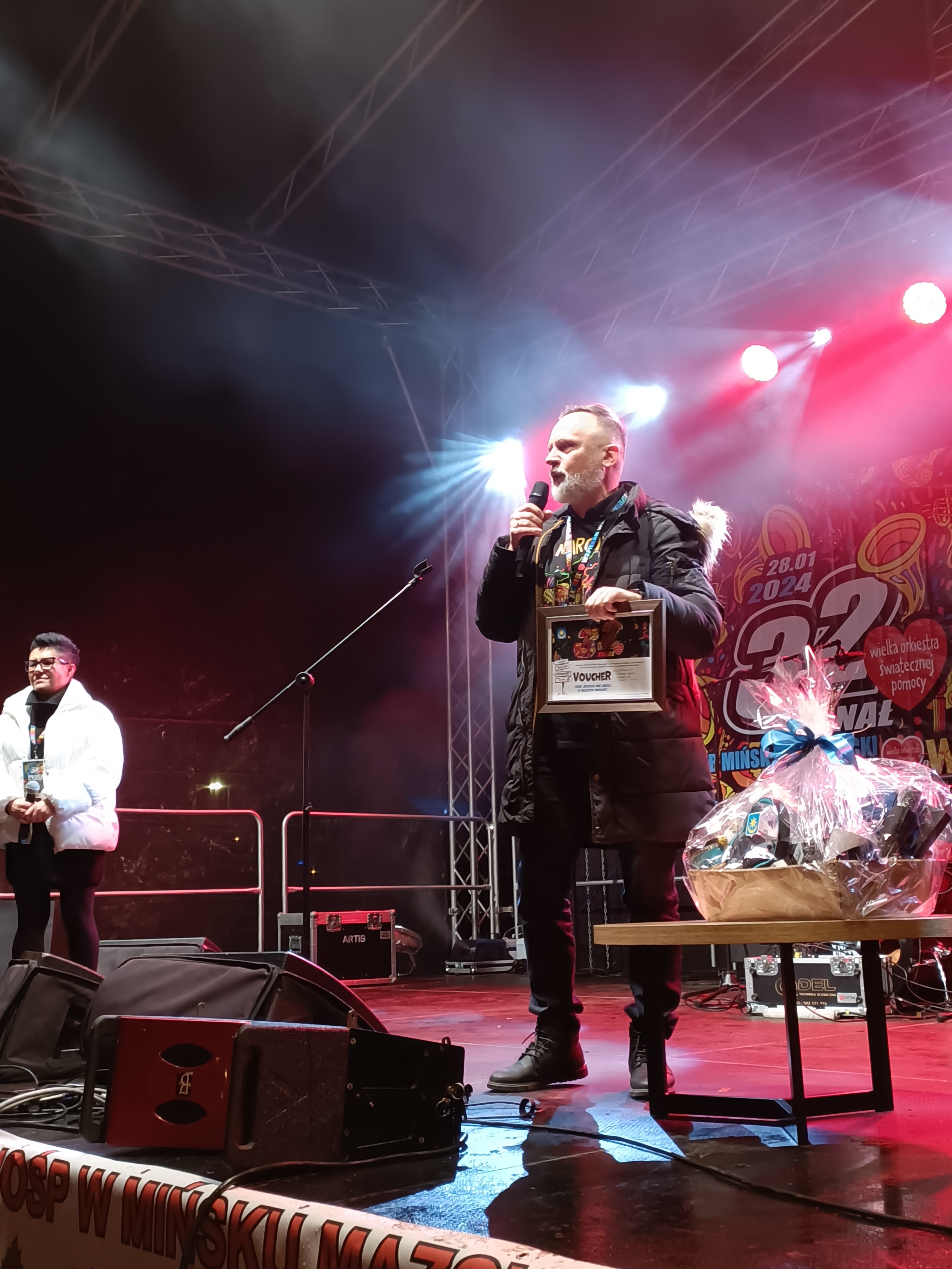 Na oświetlonej scenie kobieta z lewej, na przodzie mężczyzna przemaiwa do mikrofonu. Obok niego stolik z upominkami