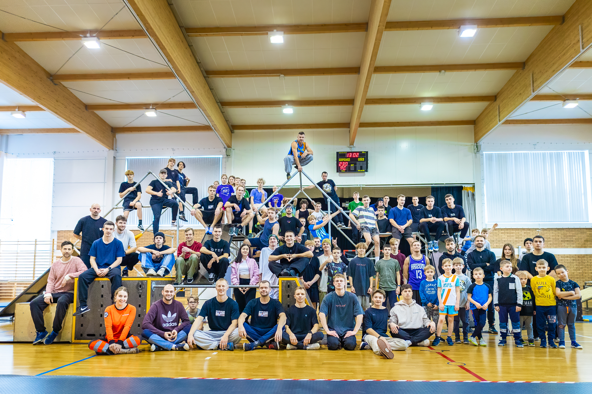 Hala sportowa. Rozstawione rusztowania do parkour. Duża grupa uczestników zawodów pozuje do zdjęcia. Częśc osób siedzi z przodu na podłodze, dalej stoją i siedzą na rusztowniach do parkour