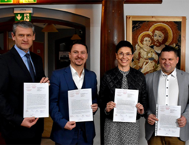 W pomieszczeniu. Pięć uśmiechniętych osób, czterech mężczyzn, po środku kobieta pozują do zdjęcią trzymając kartki w dłoniach. Za kobietą filar. W tle na ścianie obraz religijny