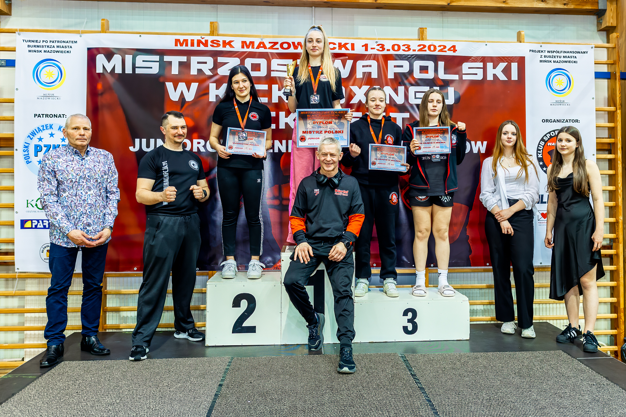 Hala sportowa. Na podium stoją osoby. Po bokach stoi po dwie osoby. Na przodzie na podium siedzi mężczyzna