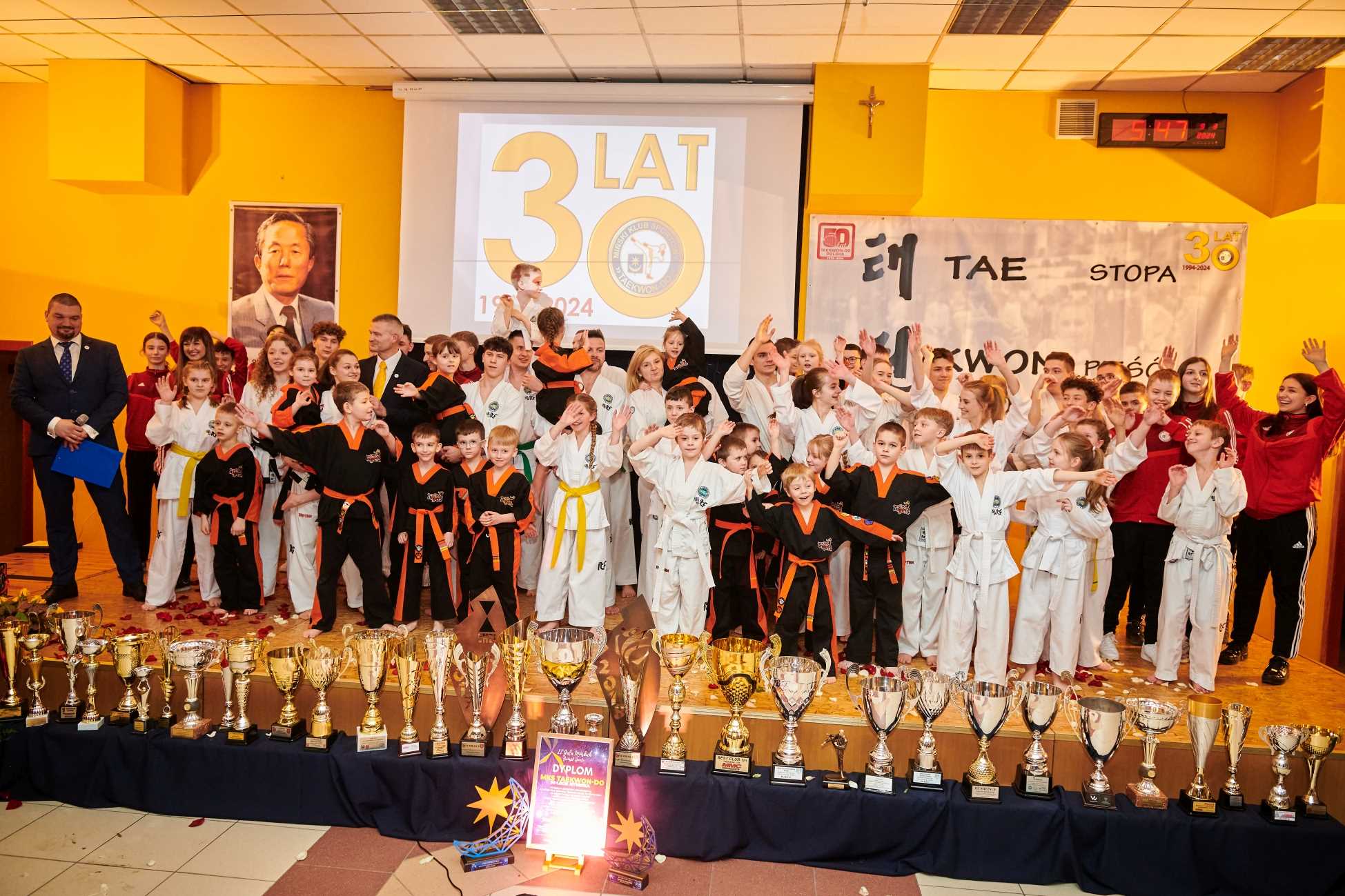 Aula. Na scenie duża grupa zawodników w białych i czarnych kimonach, nastolatki w dresach firmowych i po lewej mężczyzna w garniturze. Przed sceną puchary
