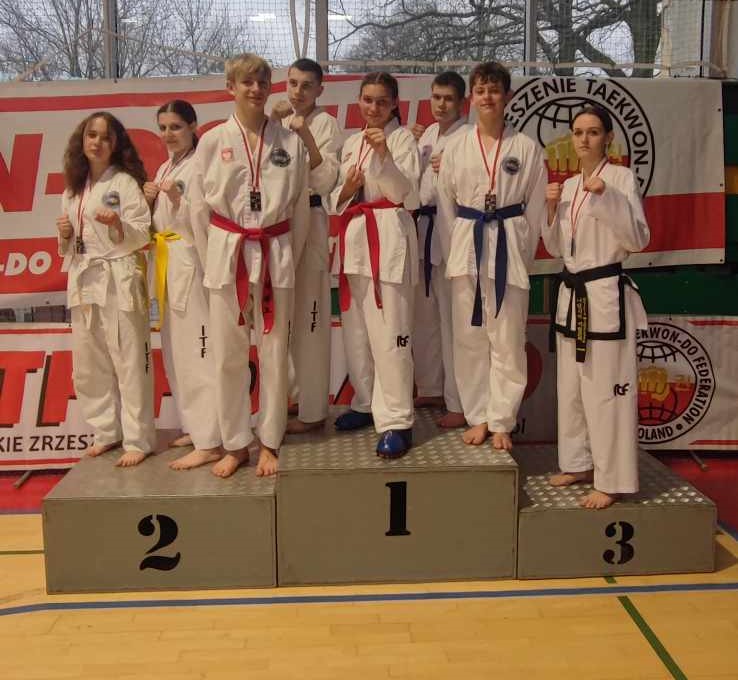 Grupa dzieci w kimonach stoi na podium. Pozują do zdjęcia w sali gimnastycznej.