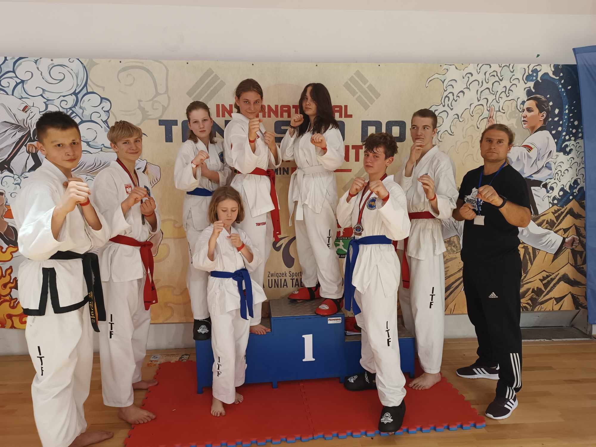 Hala sportowa. Osmioro zawodników pozuje w pozycji bojowej oraz mężczyzna
