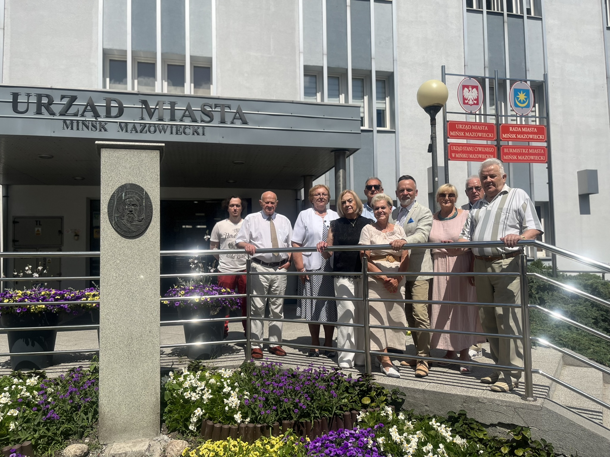 Na dworze, przed budynkiem urzędu. Dziesięc osób pozuje stojąc za metalową barierką
