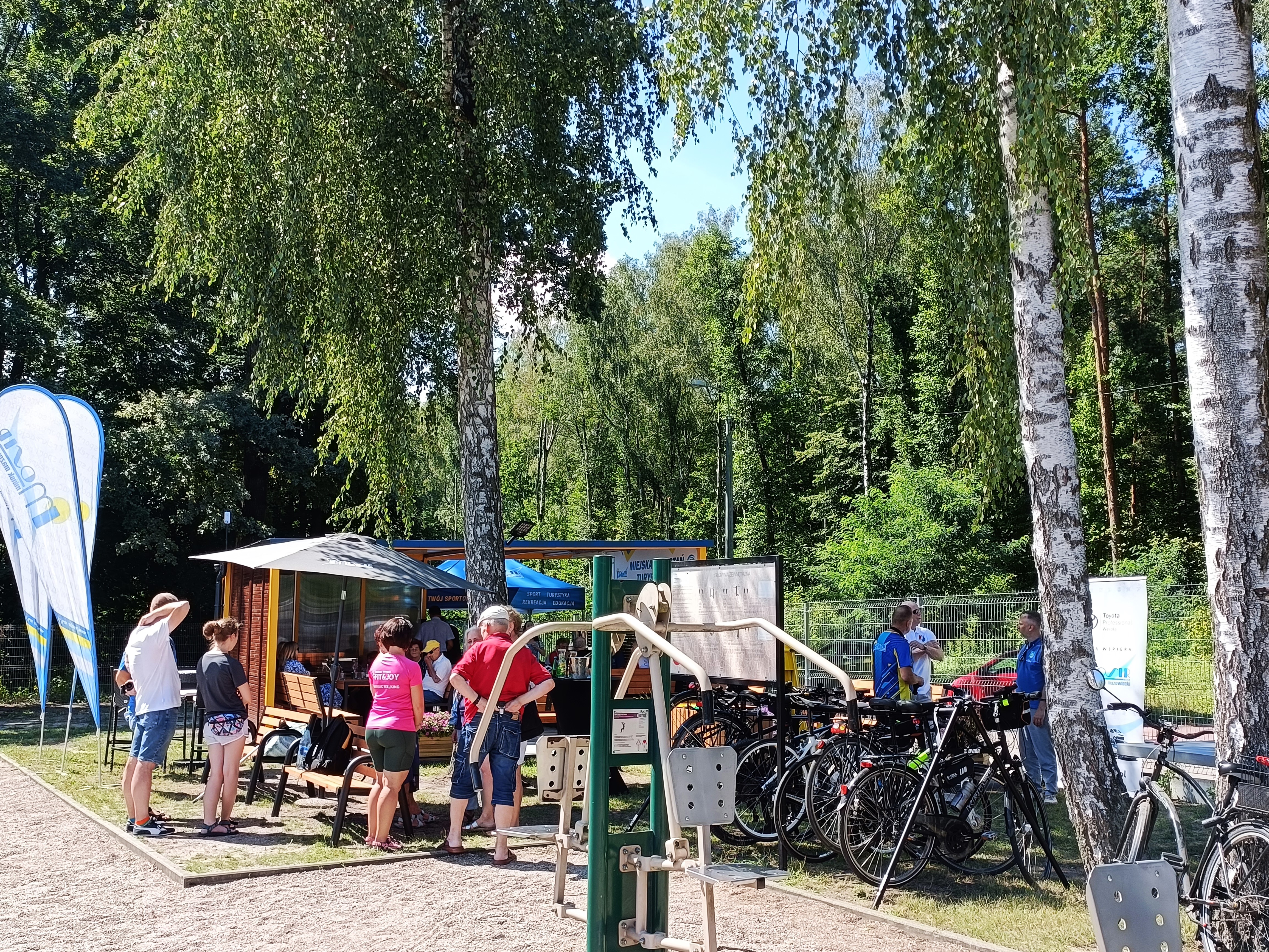 Na zewnątrz. Grupa ludzi stoi w otoczeniu zieleni. obokk rowery, w tle wiata, ławki i parasole ogrodowe.