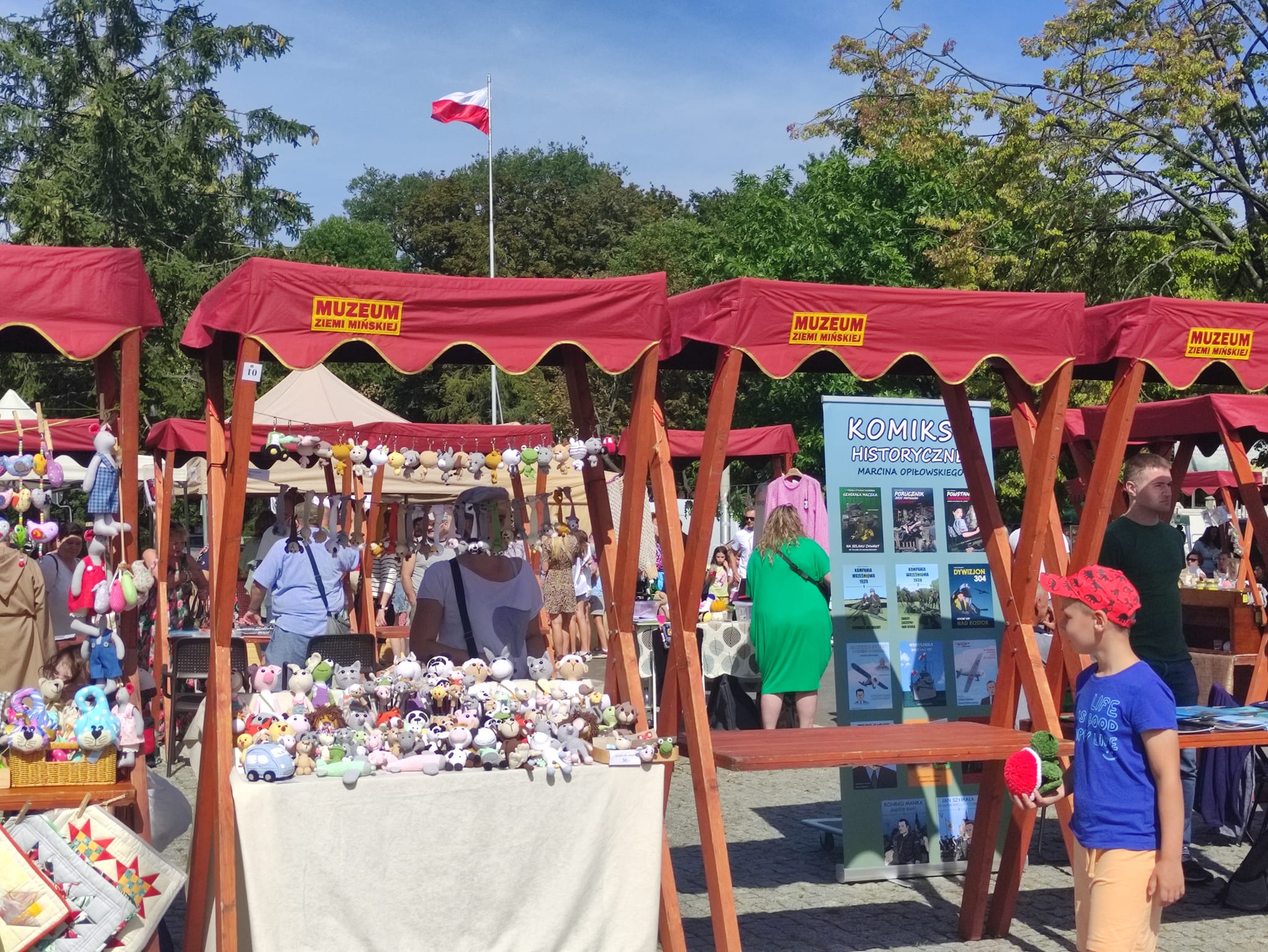 Dwór. Drewniane stragany z czerwonymi daszkami z napisem muzeum ziemi mińskiej. W tle drzewa i maszt z flagą biało-czerwoną