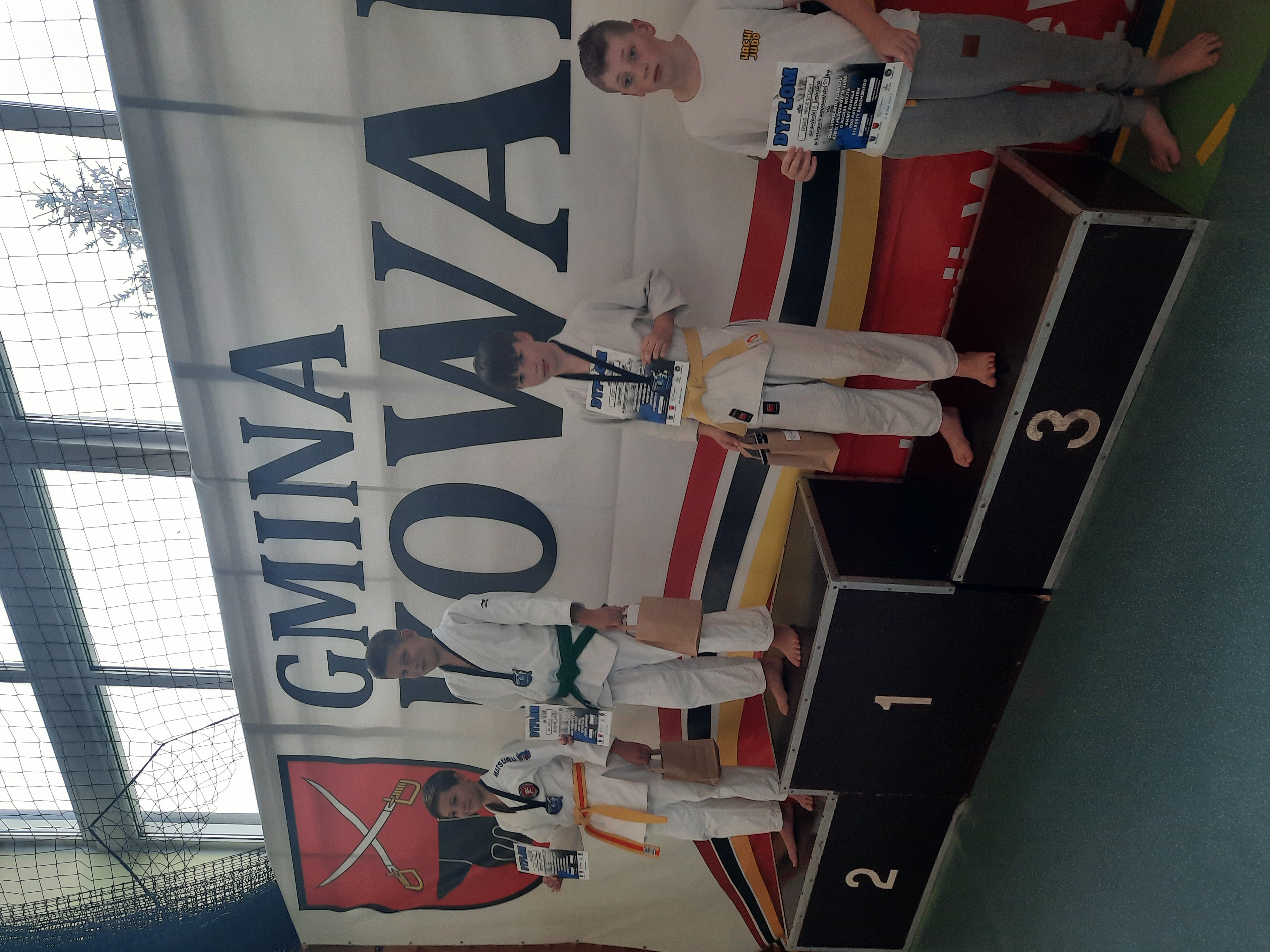 Hala sportowa. Na poszczególnych miejscach, w białych kimonach zawodnicy. Na miejscu trzecim dwie osoby