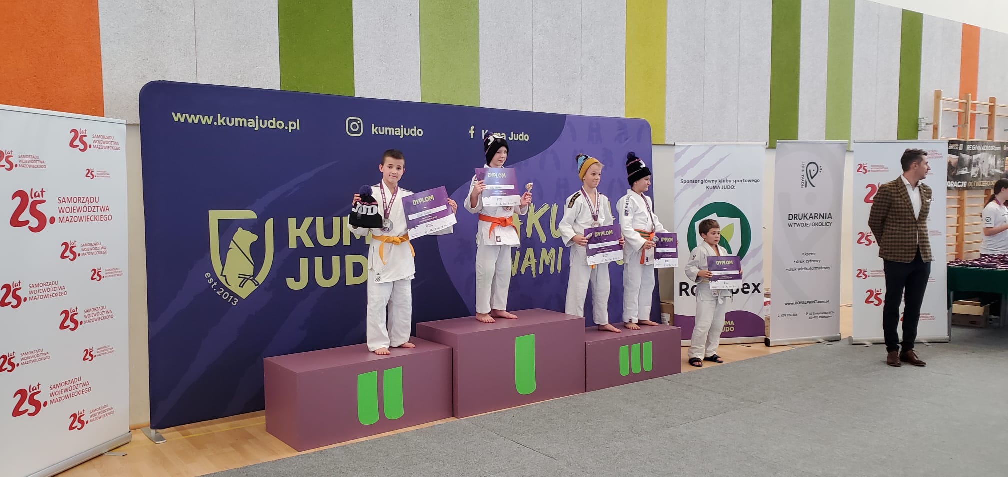 Pomieszczenie. Na wrzosowym podium czterech chłopców. Dwóch na trzecim miejscu. Po prawej piąty chłopiec. W tle banery