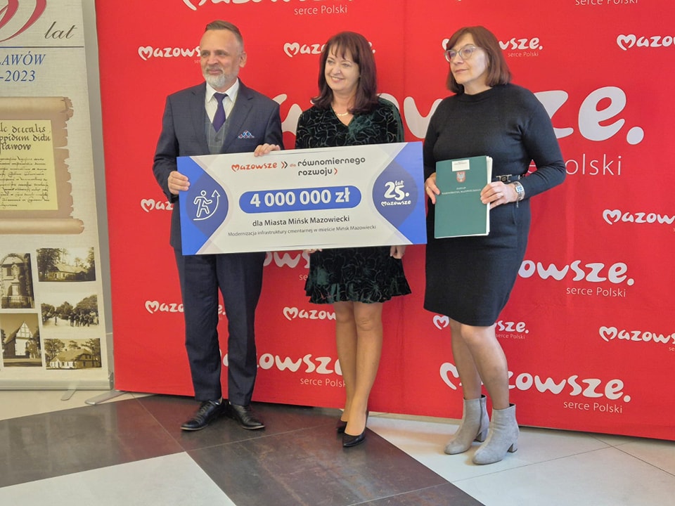 Pomieszczenie. na tle dużego, czerwonego baneru trzy osoby pozują, trzymając duże kartonowe czeki