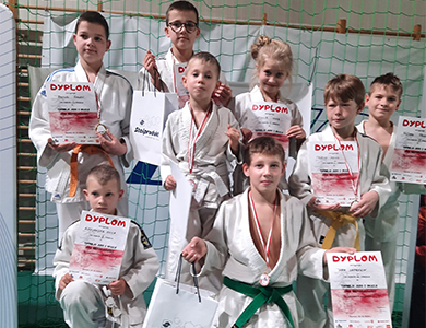Hala sportowa. Ośmioro zawdoników w białych kimonach. Trzymają dyplomy w dłoniach