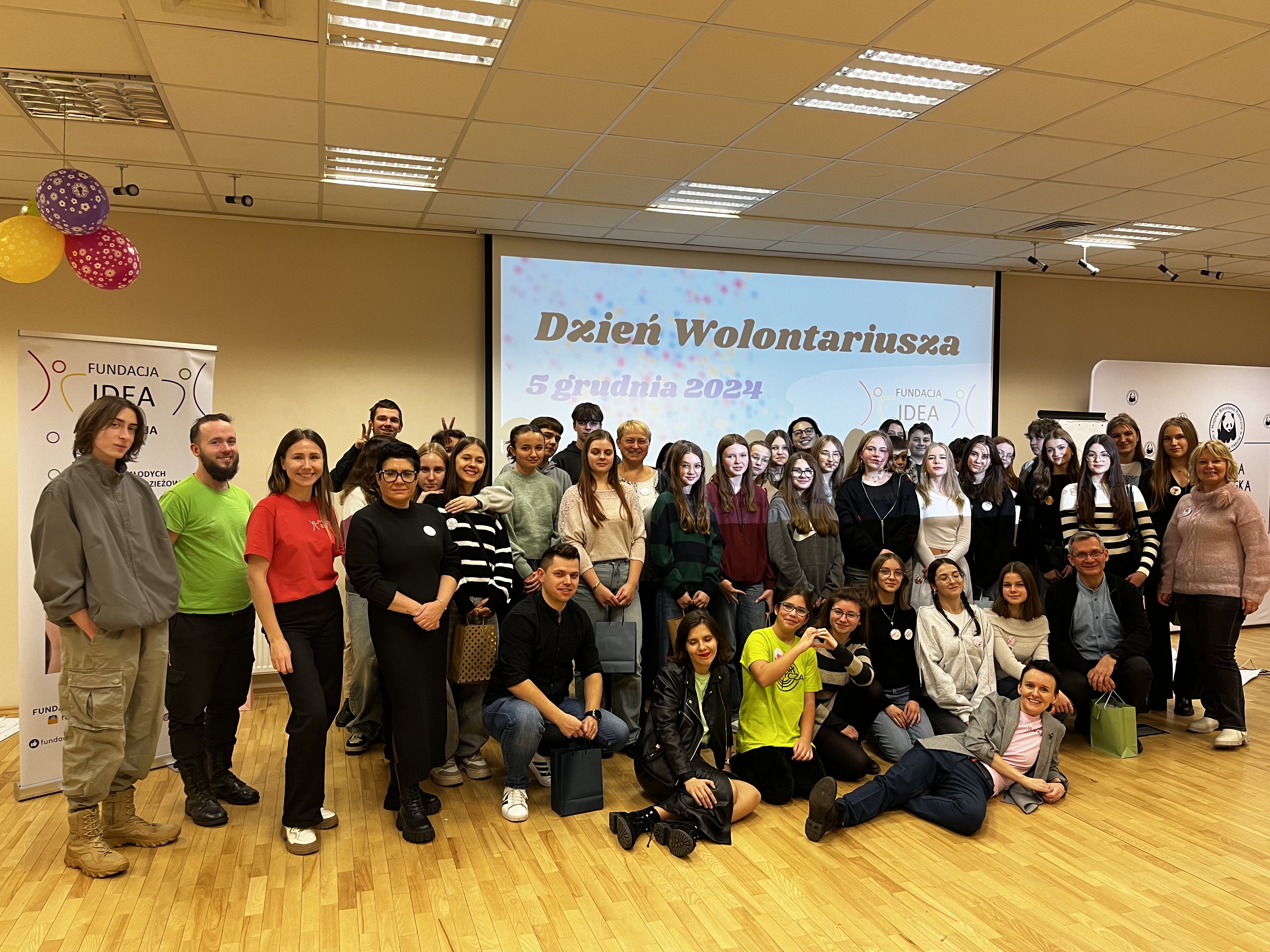 Oświetlona sala konferencyjna. Na tle dużego ekranu z tytułem Dzień Wolontariusza grupa osób