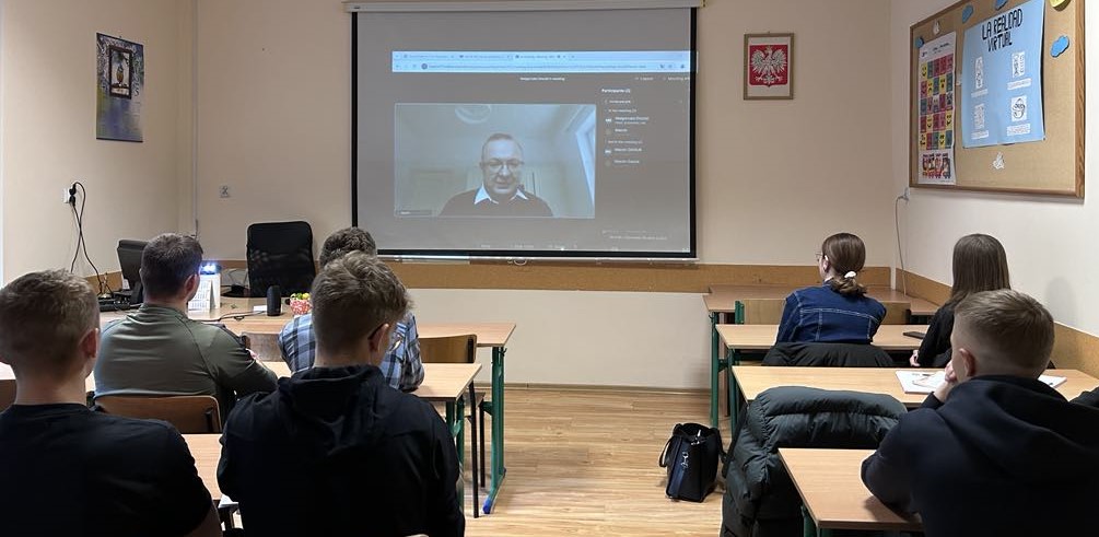 Oświetlona sala lekcyjna. uczniowie w ławkach. Na przodzie na ekranie wyświetlny mężczyzna