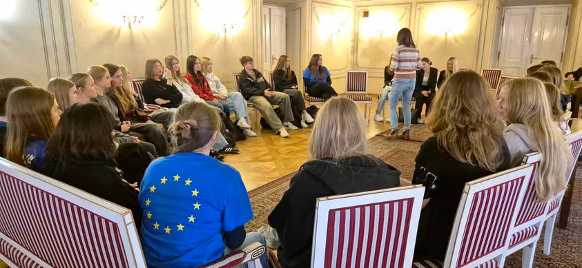 Oświetlona sala w klepce z dywanem. Naokoło na krzeslach siedzi młodzież. W środku jedna kobieta stoi tyłem