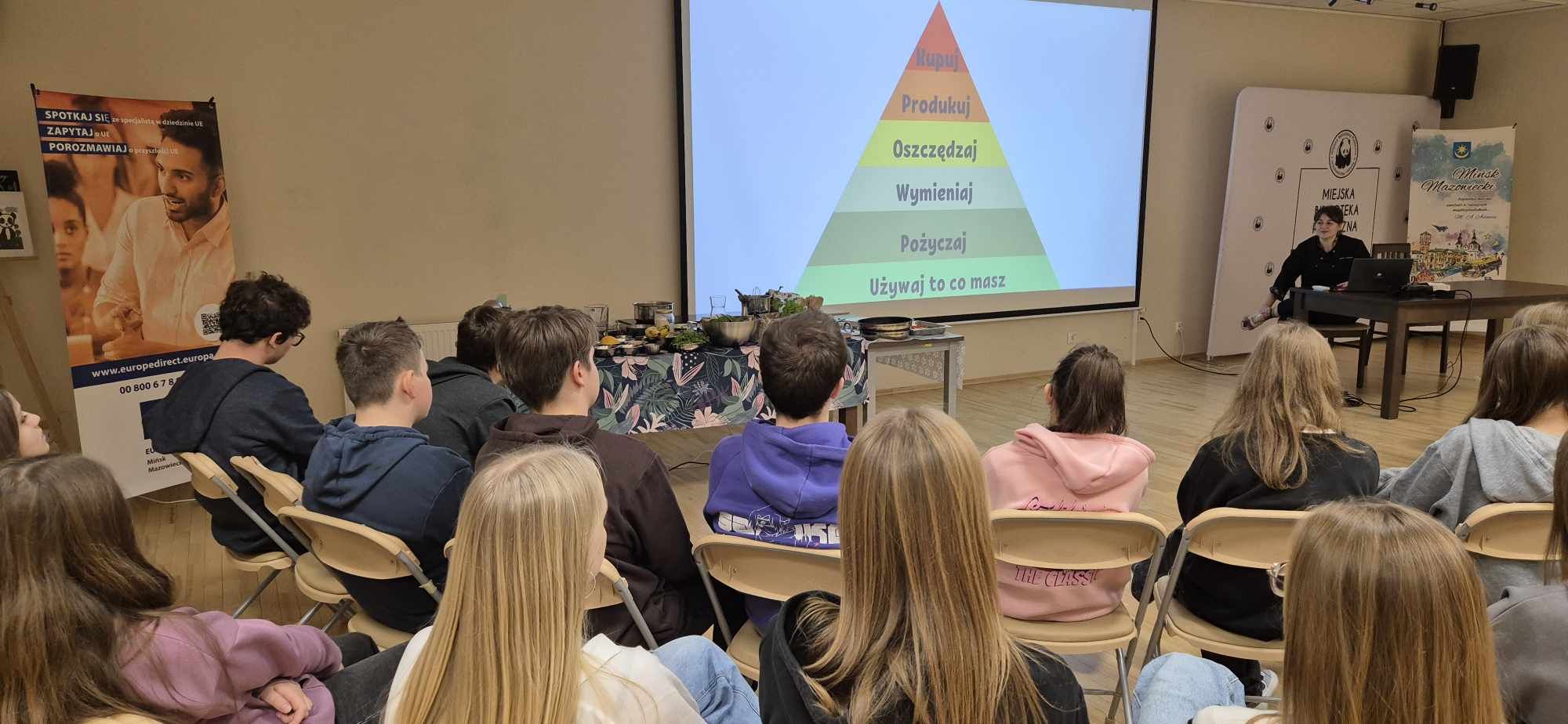 Oświetlona sala konferencyjna. Na krzesłach siedza nastolatki. Na przodzie na ekranie wyświetlana piramida żywieniowa. Po prawej przy stole na krześle siedzi kobieta