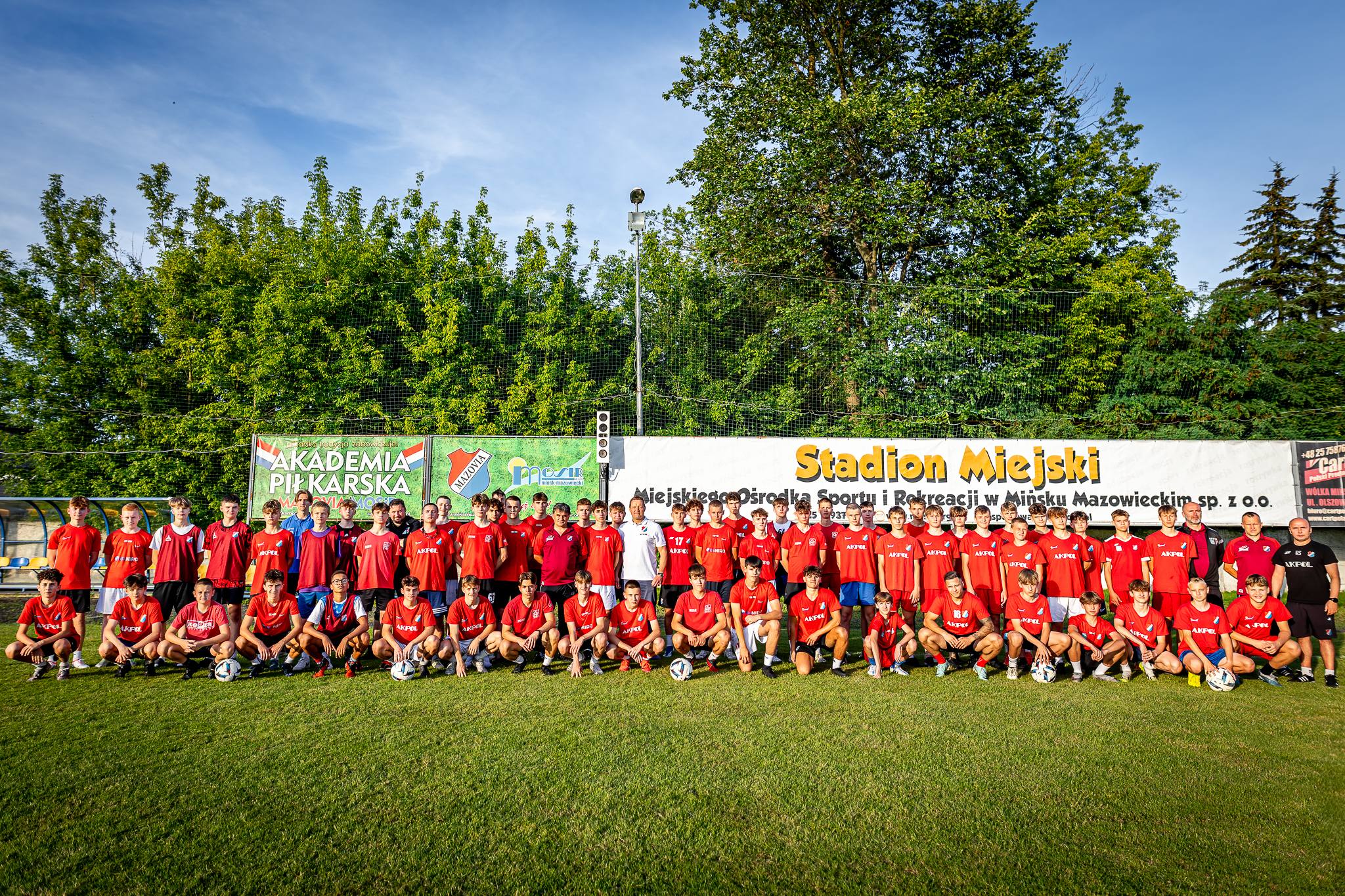liczna grupa zawodnków piłki nożnej pozuje do zdjęcia na boisku. Są ubrani w czerwone stroje sportowe.