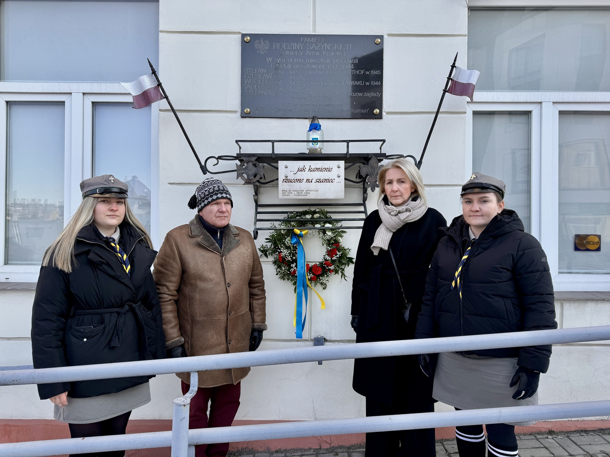 Na dworze, dzień. Przed białą ścianą budynku cztery osoby pozują do zdjęcia. Na ścianie metalowa tablica i zawieszka