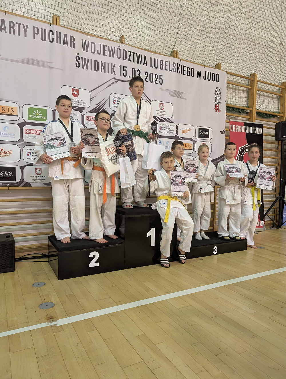 Hala sportowa. Na podium grupa zwycięzców w białych kimonach z dyplomami w dłoniach. W tle baner zawieszony na drewnianych drabinkach
