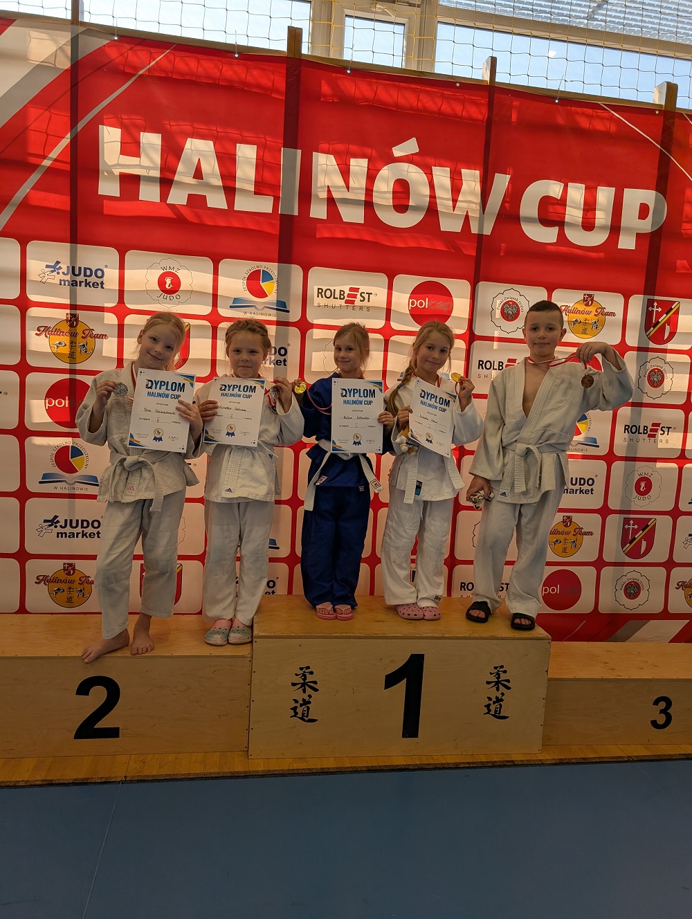 Oświetlona hala sportowa, dzień. Na drewnianym podium piątka dzieci w kimonach. Cztery dziewczynki trzymają dyplomy. Chłopiec podnosi medal. W tle czerwony baner z logami