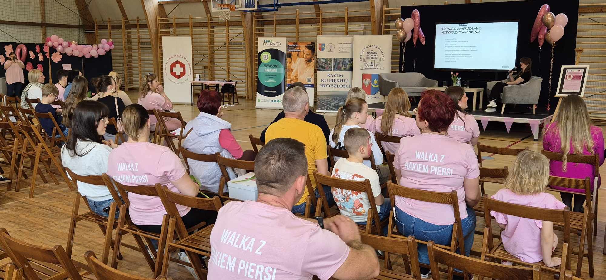 Oświetlona hala sportowa. na krzesłach sluchacze. Na przodzie scena z dwiema kanapami i stolikiem. Po lewej banery