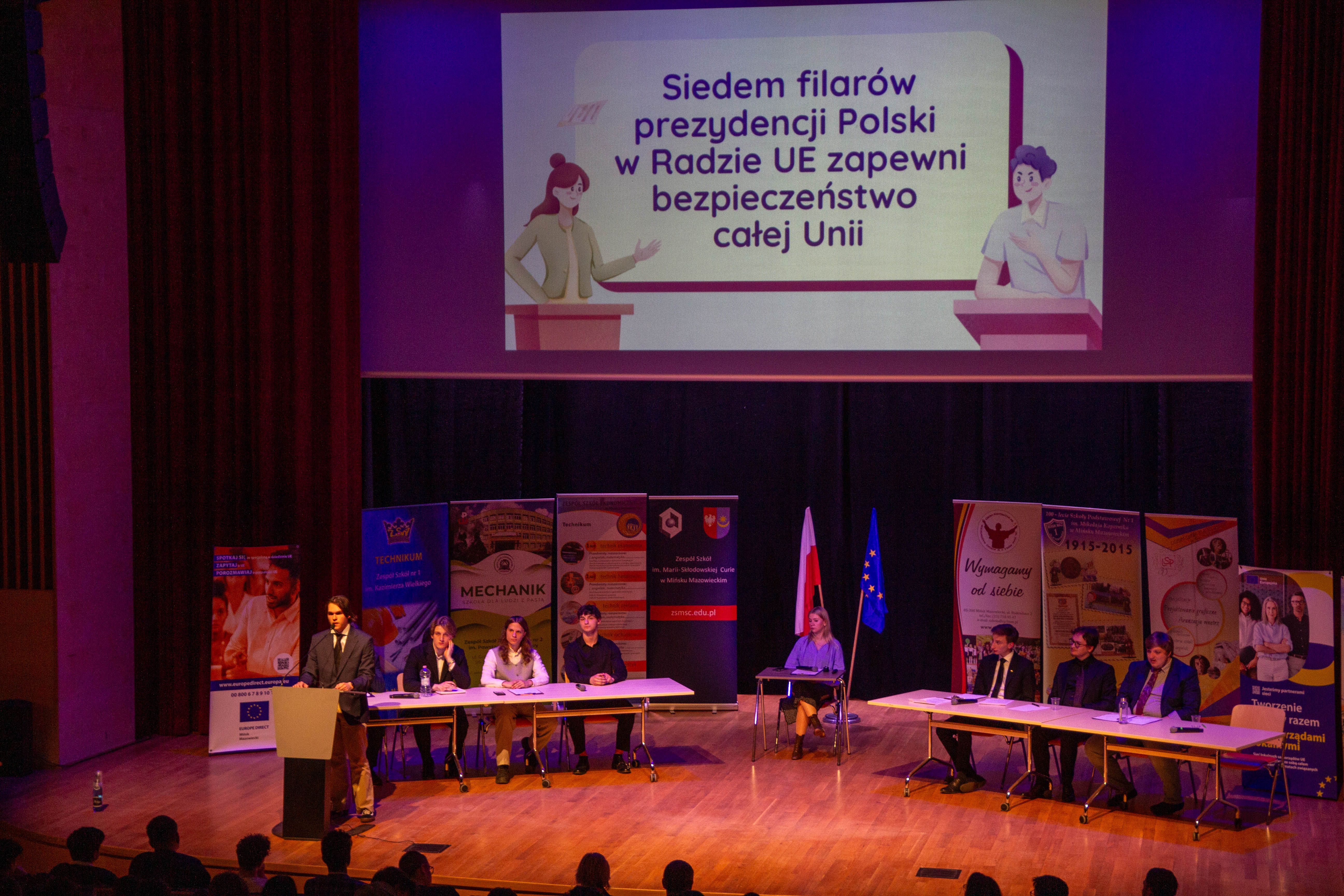 Aula, widok na scenę. Dwa długie stoły, po środku mały. Przy nich osoby. W tle stoją banery. Nad sceną wisi ekran z treścią