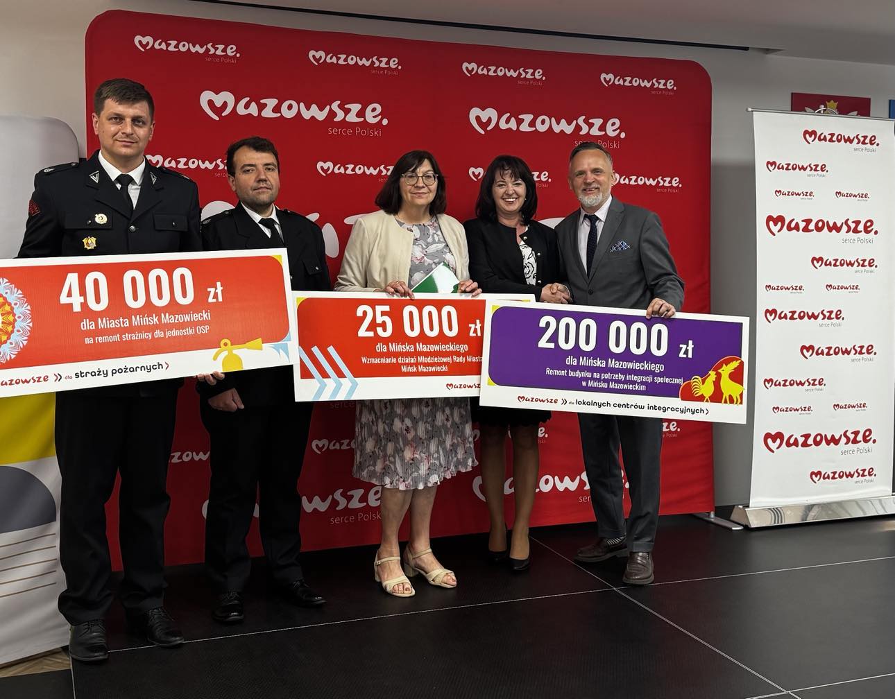 Oświetlone pomieszczenie. Pięć osób pozuje trzymając duże banery. Na nich kwoty. W tle czerwony duży baner z białymi napisami Mazowsze