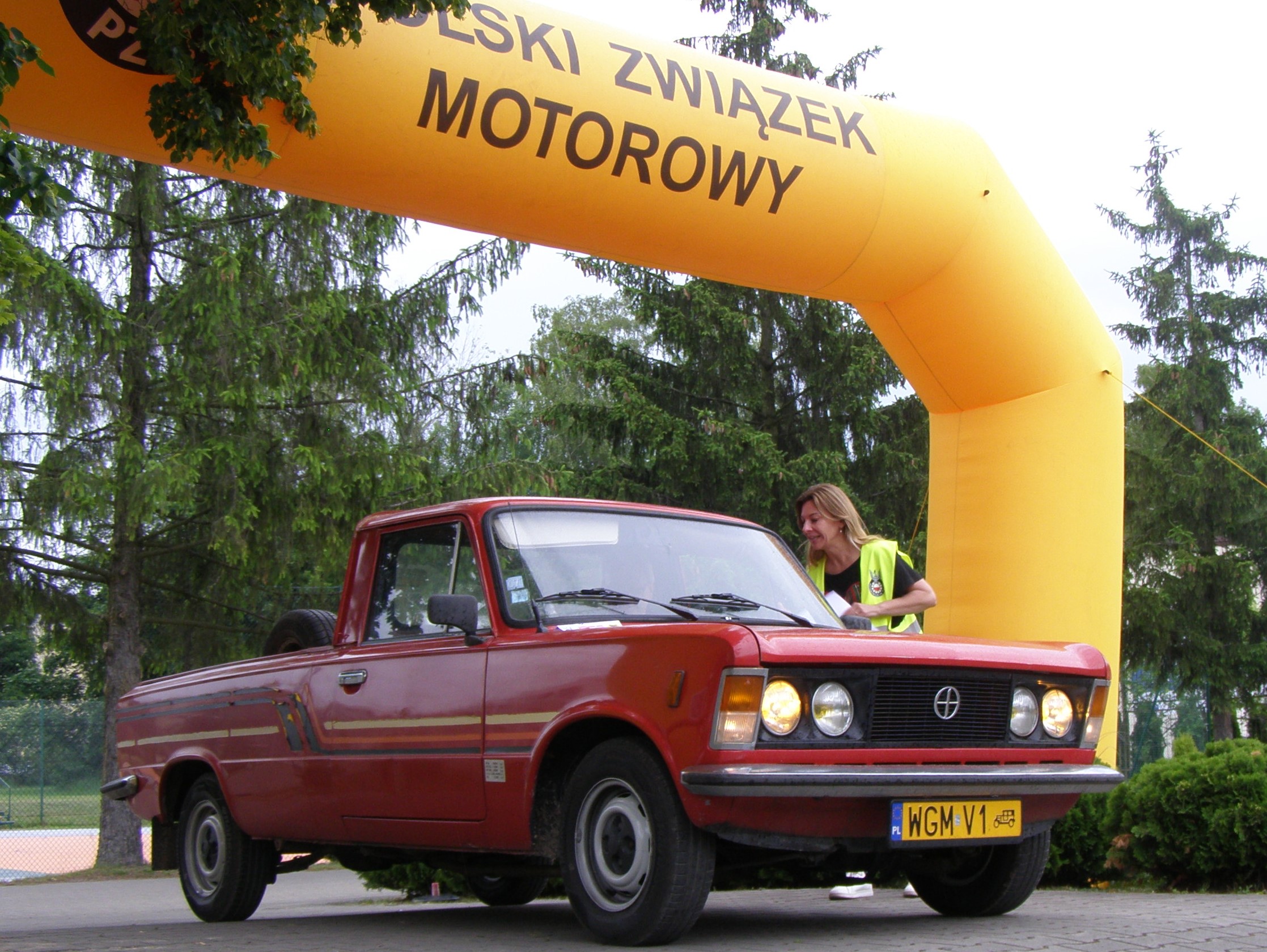 Dzień. Czerwony zabytkowy samochód stoi na linii mety. Nad nim żółtym balon. W tle drzewa