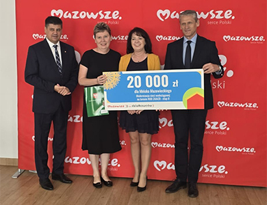 W jasnym pomieszczeniu, na drewnianej klepce dwóch mężcyzn i dwie kobiety. Trzymaja baner z kwota 20000. W ich tle czerwony duzy baner z napisem Mazowsze