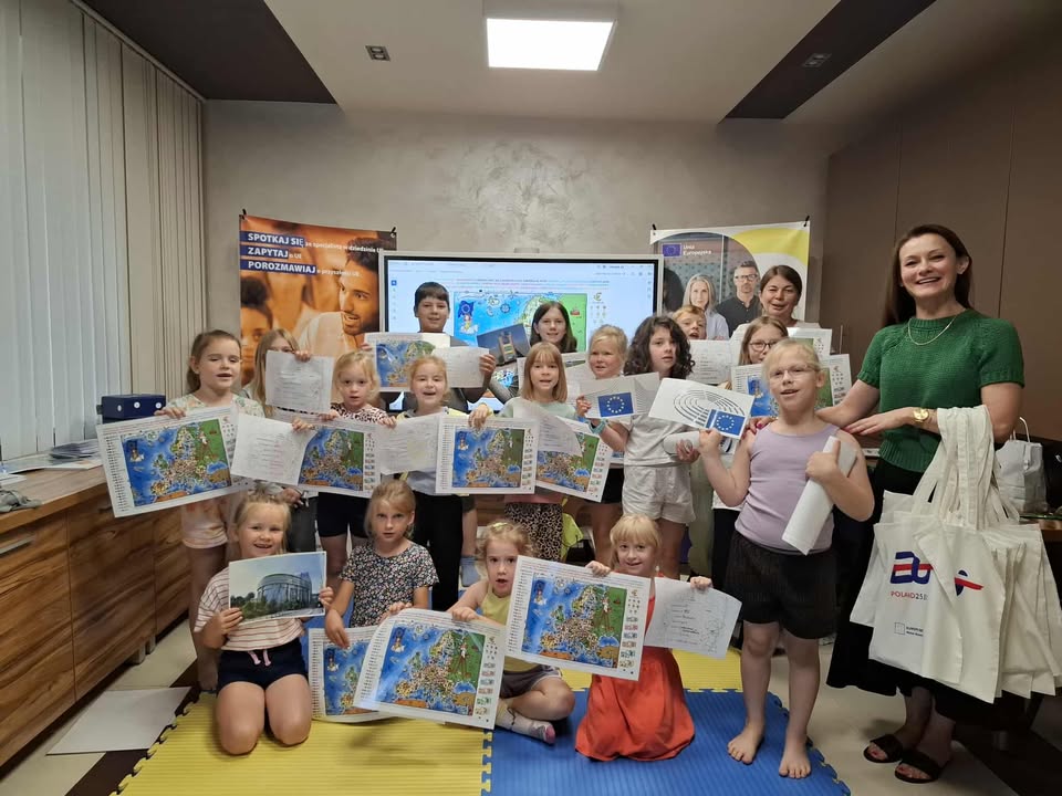 Jasne pomieszczenie. Grupa dzieci z mapami w dłoniach. Po prawej usmiechnięta kobieta w zielonym swetrze i zawieszonymi na przedramieniu jasnymi lnianymi torbami